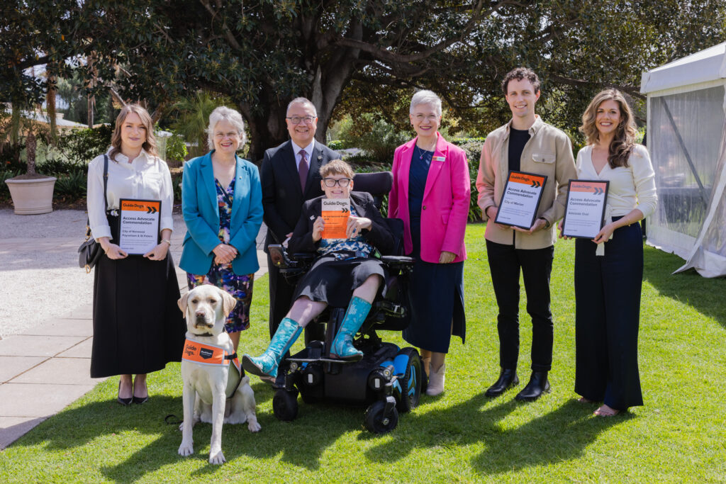 Seven people, one in a wheelchair, stand in a row outdoors holding certificates. A guide dog wearing an orange vest sits in front of the group.