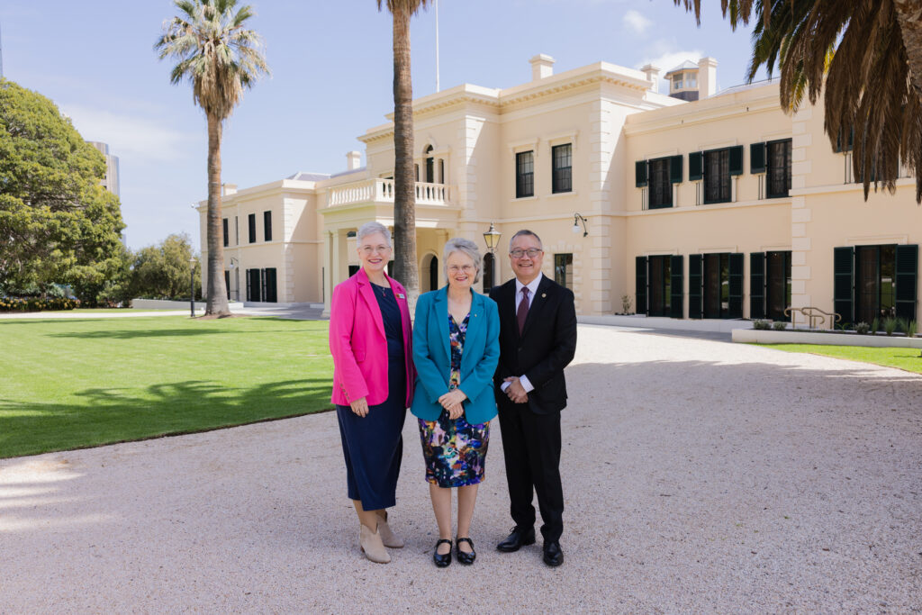 Three people stand smiling in front of a large beige building with columns, surrounded by green lawn, trees, and blue sky.