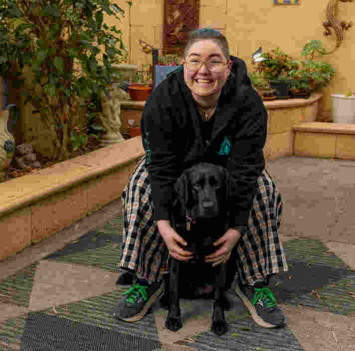 A person with blue hair and glasses smiles while crouching behind a black dog in a patio garden area with potted plants and decorative wall art.