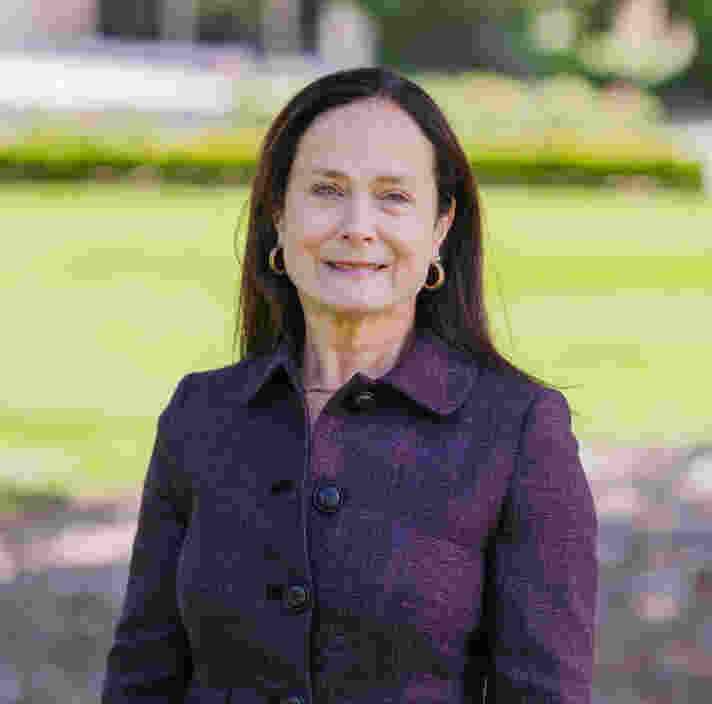 A woman with straight dark hair, wearing a purple blazer, stands outdoors on a sunny day with grass and trees in the background.