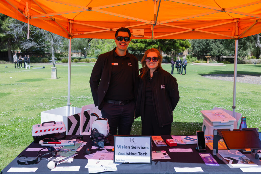 Two people stand behind a table displaying assistive technology devices and brochures under an orange canopy labeled Vision Services Assistive Tech in an outdoor setting.