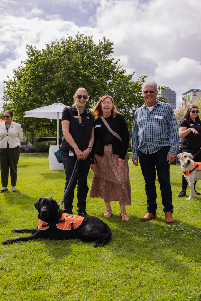 Three adults stand on grass with a black guide dog wearing an orange harness lying in front, while another guide dog and people are visible in the background.