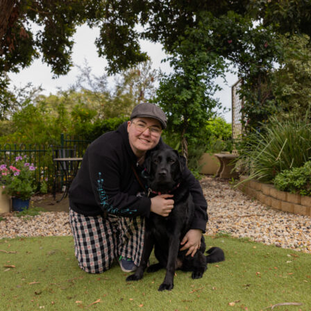 A person kneels on grass in a garden, smiling and hugging a black dog. There are trees, plants, and potted flowers in the background.