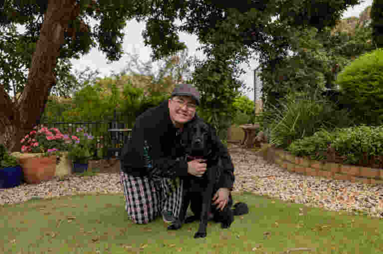 A person kneels on grass in a garden, smiling and hugging a black dog. There are trees, plants, and potted flowers in the background.