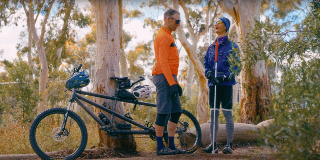 Two people in outdoor sportswear stand next to a tandem bicycle in a wooded area. One person holds walking poles and wears glasses; both are wearing helmets and gloves.