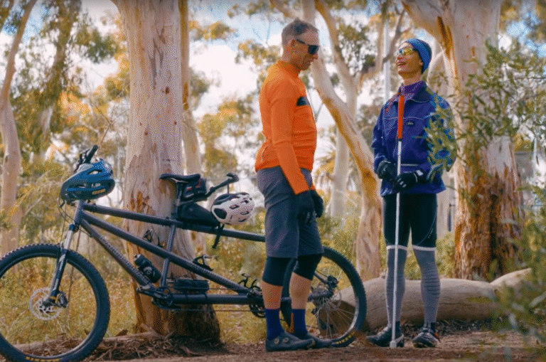 Two people in outdoor sportswear stand next to a tandem bicycle in a wooded area. One person holds walking poles and wears glasses; both are wearing helmets and gloves.