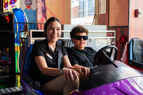 A woman and a boy wearing sunglasses sit together in a bumper car at an indoor amusement arcade, both smiling at the camera.