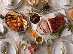 A holiday table setting with sliced roast pork, roasted vegetables, sauces, olives, orange slices, and gift box, with plates, cutlery, and glasses arranged neatly.