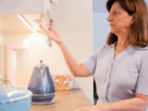A woman stands in a kitchen holding her hand under a cabinet light, next to an electric kettle on the countertop.