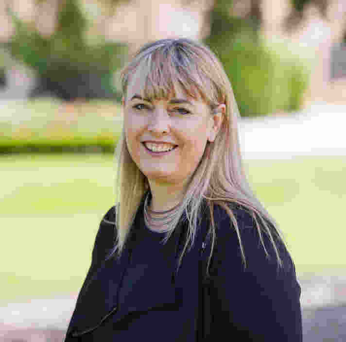 A woman with long blonde hair and bangs, wearing a black top, stands outdoors smiling with greenery and trees in the background.