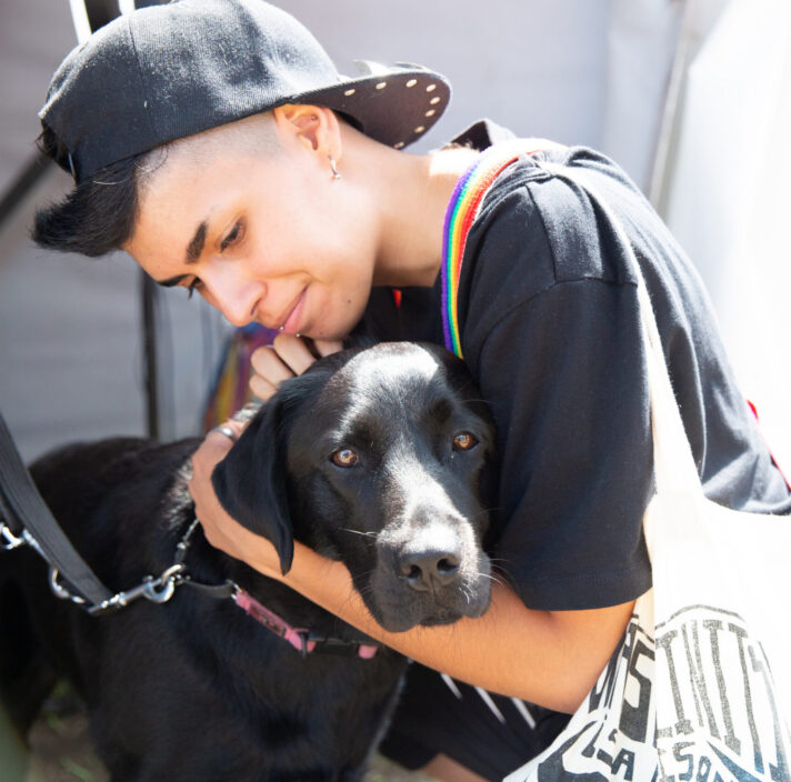 Person in a black shirt and cap hugs a black dog closely. The scene is well-lit, and the person carries a white tote bag with black text.