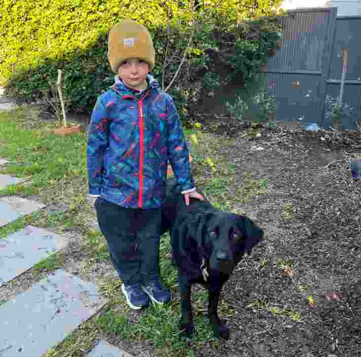 A young boy in a blue patterned jacket and yellow beanie stands next to a black dog on a garden path with greenery and a fence in the background.
