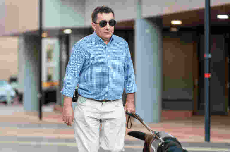 A man wearing sunglasses walks with a guide dog on a city sidewalk.