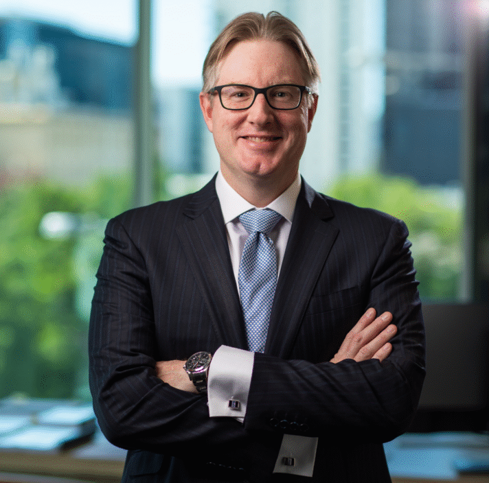A man in a suit and tie stands with arms crossed in an office with large windows and a city view in the background.