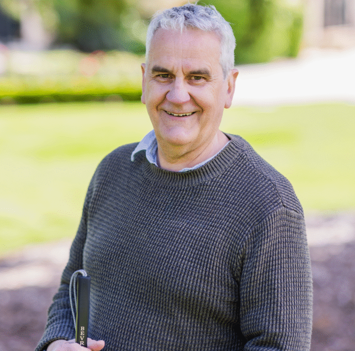 An older man with gray hair, wearing a brown sweater, stands outdoors holding a black-handled cane, smiling at the camera with a grassy background.