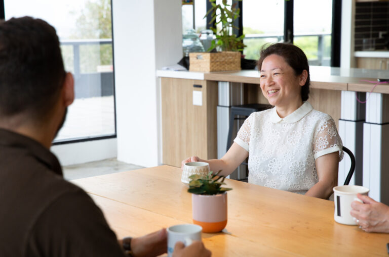 Three people sit at a wooden table in a modern kitchen, holding mugs and having a conversation.