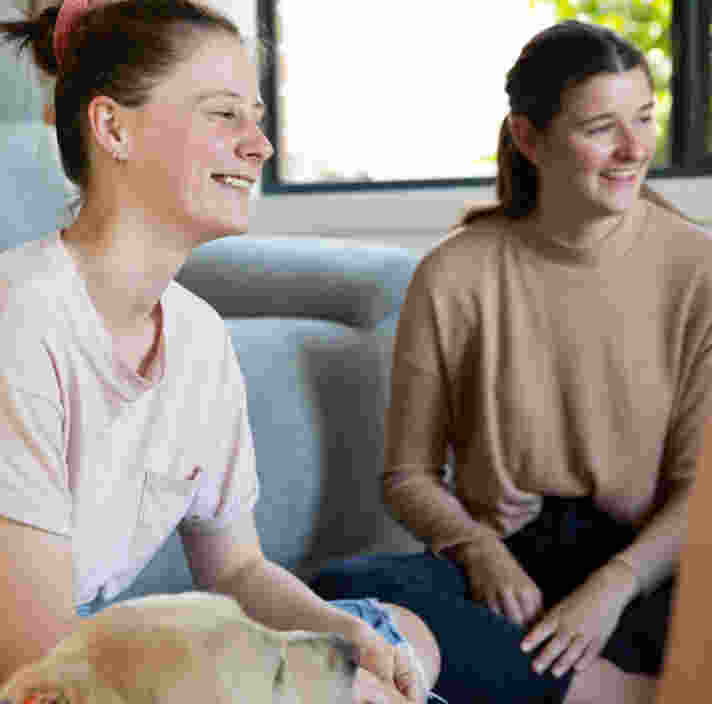 Two young women sit indoors, smiling and talking together. A dogs head is visible in the foreground. A third person is partially visible on the right.