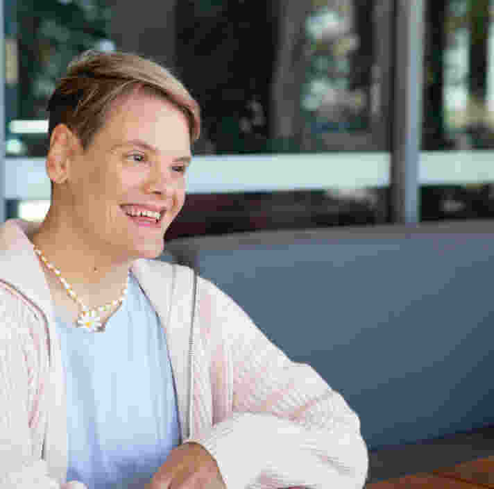 A person with short hair sits at a wooden table outdoors, smiling and wearing a light sweater, blue top, and a beaded necklace.