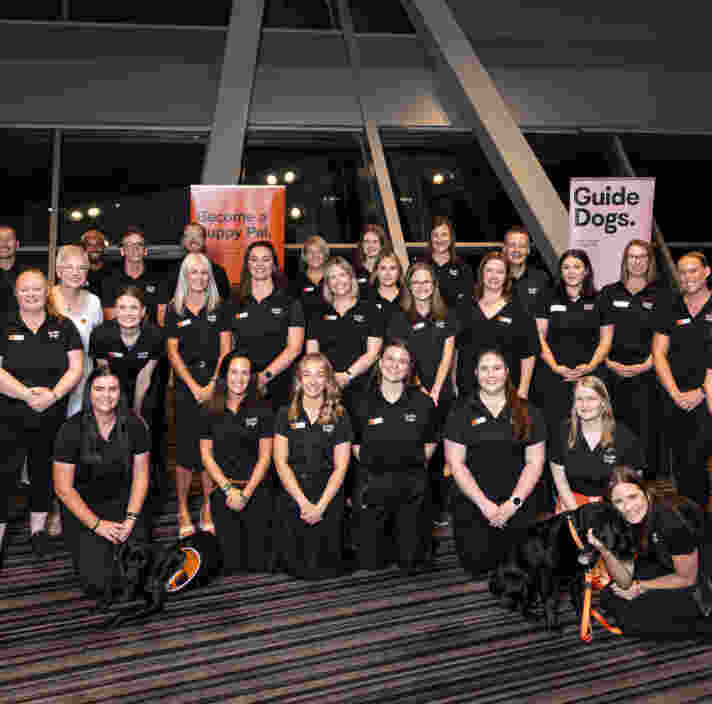 A large group of Guide Dogs Staff in black uniforms pose indoors in front of Become a Puppy Pal and Guide Dogs signs, with two guide dogs at the front.