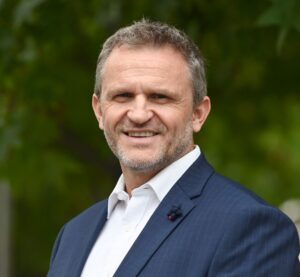 Guide Dogs SA/NT CEO Daniel Cox in a blue suit and white shirt smiles at the camera, standing outdoors with green foliage in the background.