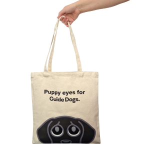 A hand holds the Tote Bag - Puppy Eyes, a beige tote bag with a cartoon dog face and "Puppy eyes for Guide Dogs" text.