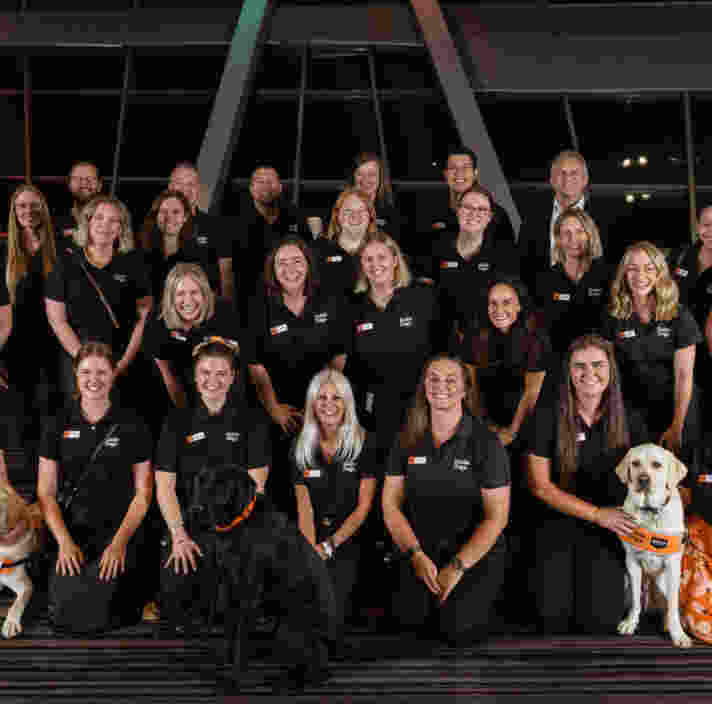 A group of people in matching black shirts pose indoors for a group photo, with three guide dogs seated in front.