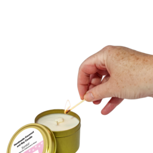 A hand lights a white coconut and soy Candle Tin - Gold with a pink label that reads Handmade Coconut and Soy Candle, Santal.