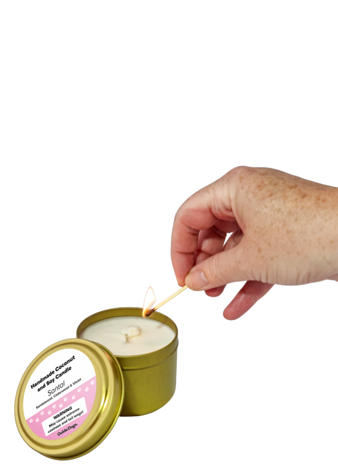 A hand lights a white coconut and soy Candle Tin - Gold with a pink label that reads Handmade Coconut and Soy Candle, Santal.