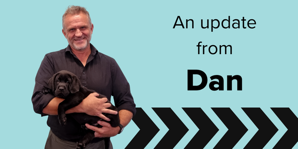 Guide Dogs SANT CEO Dan holding a black puppy stands in front of a blue background with the text An update from Dan and black chevron graphics at the bottom.