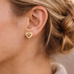 Close-up of a person with light brown hair in a low bun, wearing Earrings - Heart: gold earrings shaped like hearts featuring a cut-out paw print design.