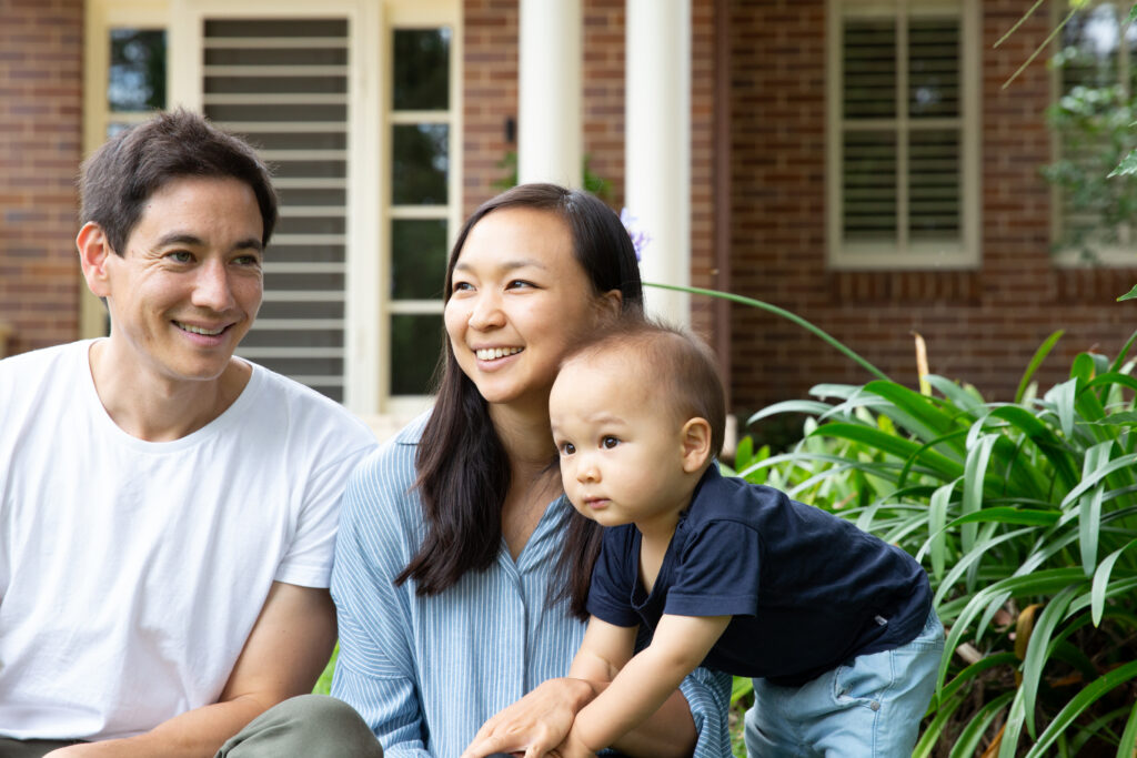A young family with a toddler child sitting outside of a home in a garden.