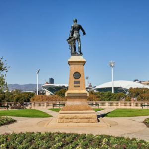 A bronze statue of a Colonel Light stands on a tall stone pedestal in a landscaped park, with Adelaide Oval and floodlights visible in the background under a clear blue sky.