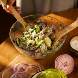 A person mixes a bowl of salad with sliced cucumbers, red onions, and herbs. Sliced red onions and a bowl of salt are on the wooden table nearby.