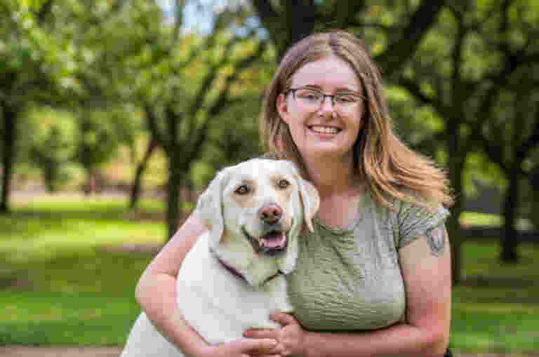 Woman with glasses smiling and sitting outdoors, holding a white dog with a purple collar on a wooden bench, with green trees in the background.