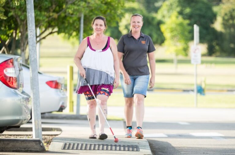 A person crossing the road with their white cane while a Guide Dog's staff member walks beside them. They are both looking at the camera smiling.