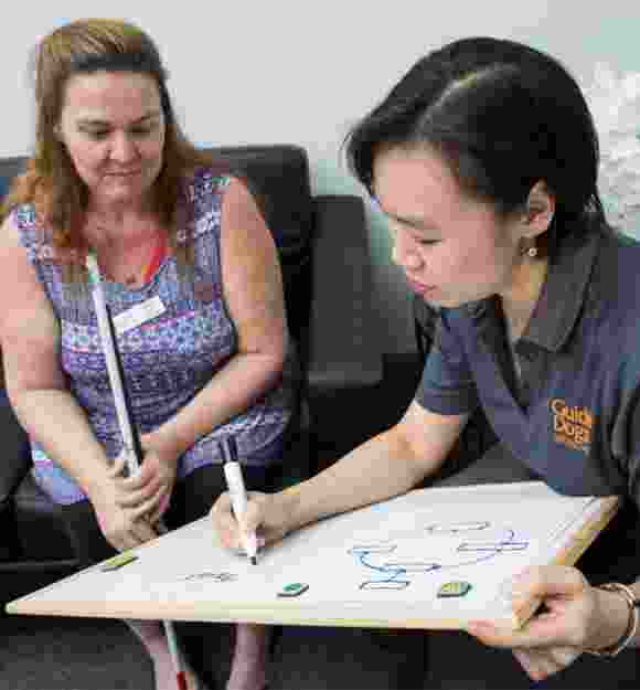 A person sitting with a Guide Dog's staff member. The staff member is writing on a small white board on her laptop while the person is looking at the board. The person is holding a white cane in their hands.