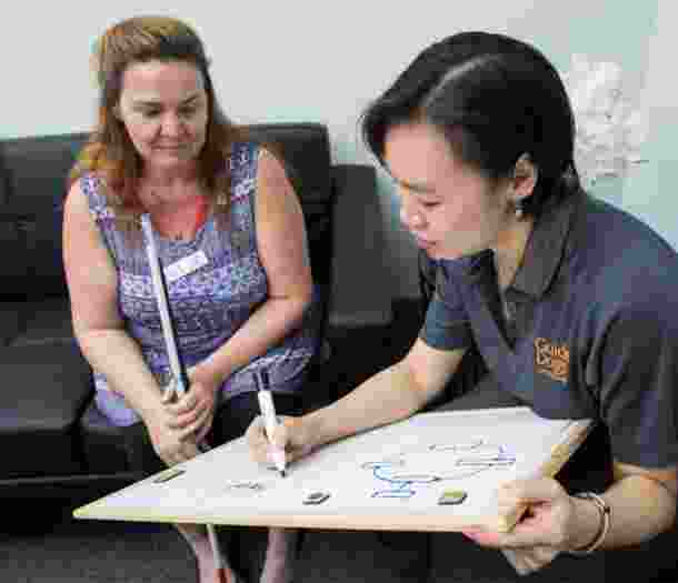 A person sitting with a Guide Dog's staff member. The staff member is writing on a small white board on her laptop while the person is looking at the board. The person is holding a white cane in their hands.