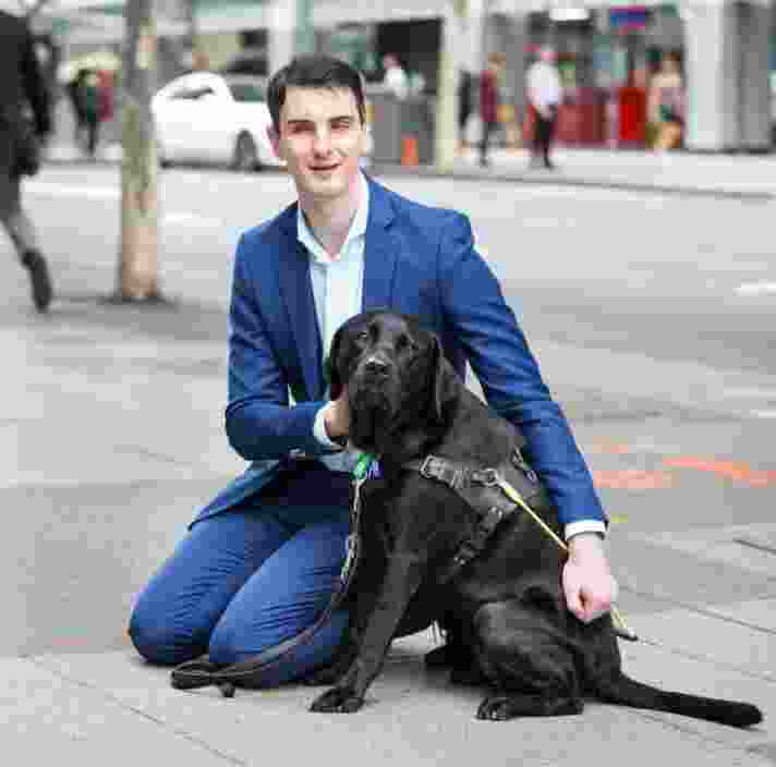 Person kneeling down outside next to a black Guide Dog in harness. The person and Guide Dog are looking at the camera.