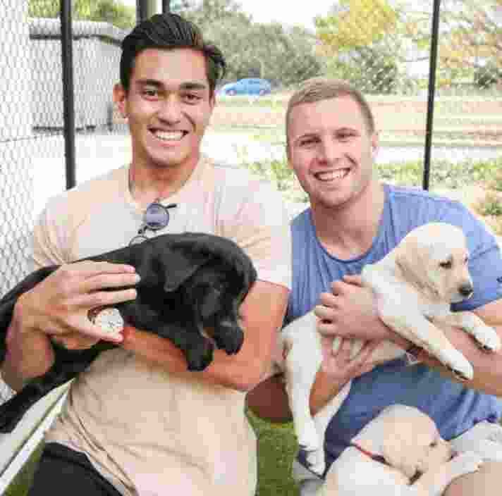 Two people seated outside holding eight week old labrador puppies. One of the puppies is yellow and the other is black. Both people are looking at the camera smiling.