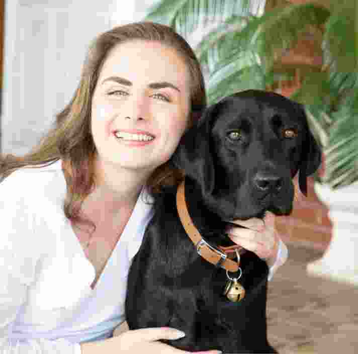 A person looking at the camera smiling with their arm around the neck of a black labrador dog.