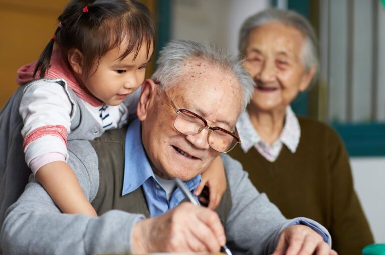 An older man writing while a young girl puts her arms around his shoulders, hugging him. The man is smiling and there is an older woman in the background who is not in focus but is also smiling.