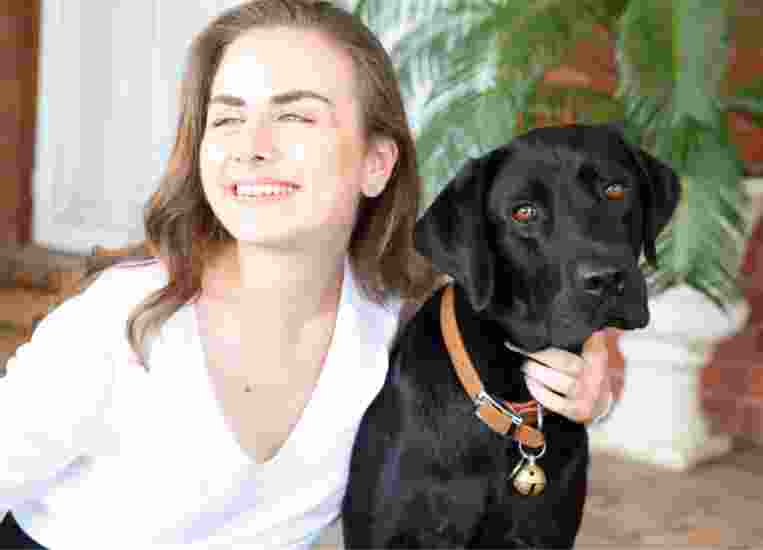 A person looking to the left of the camera smiling with their arm around the neck of a black labrador dog.