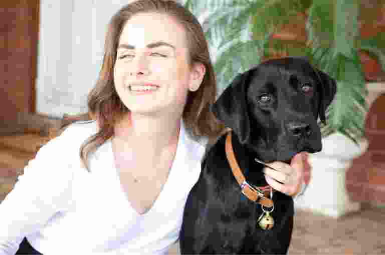 A person looking to the left of the camera smiling with their arm around the neck of a black labrador dog.