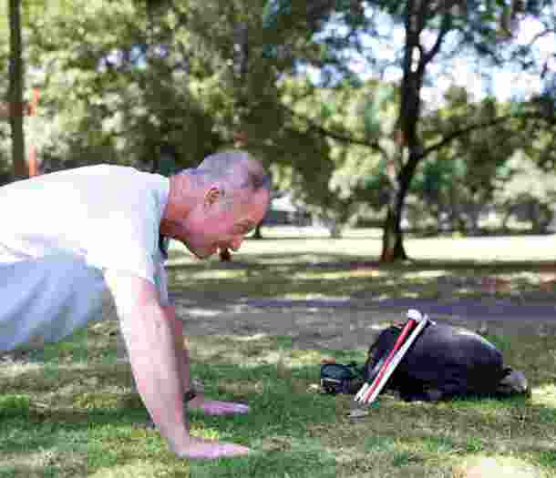 A person doing push ups outside in the park. There is a black bag near them with a folded white cane leaning up against the bag.