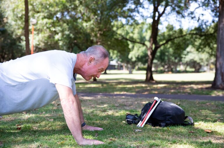A person doing push ups outside in the park. There is a black bag near them with a folded white cane leaning up against the bag.