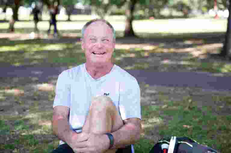 A person sitting outside on the grass with a smile on their face looking at the camera. They have one leg bent with their arm wrapped around their knee and a white cane next to them.