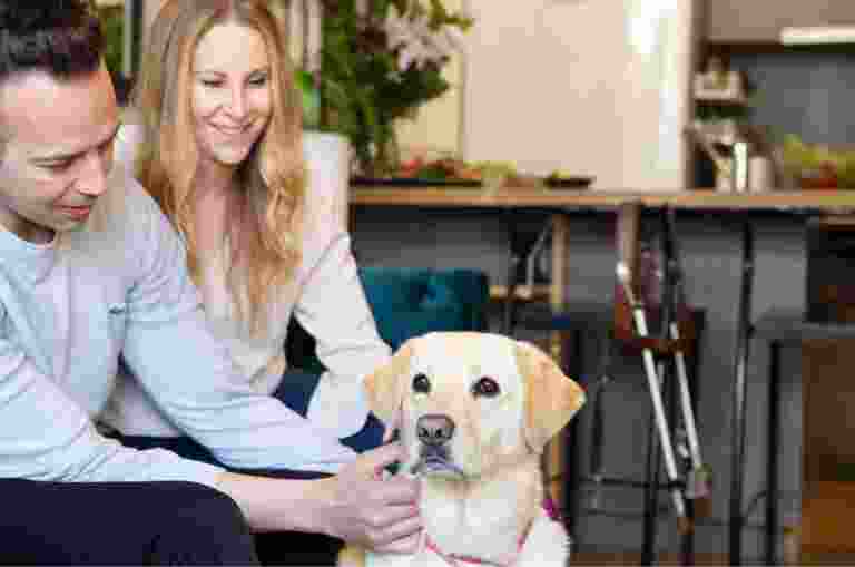 Two people sitting on their couch patting their yellow labrador dog.
