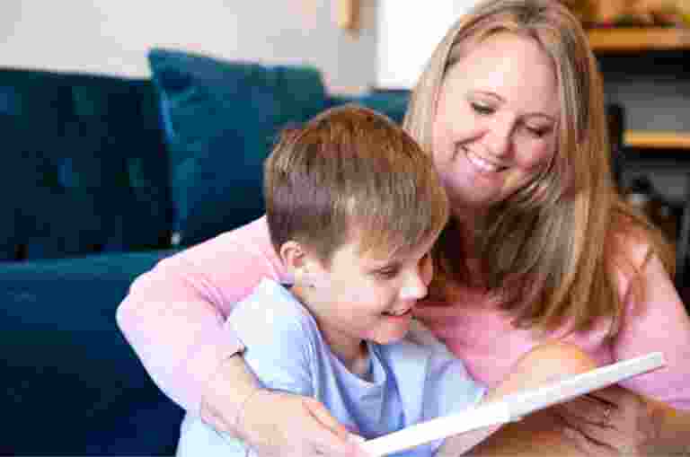 Young child sitting on the ground with their mother next to them. They are both smiling and looking at an Ipad.