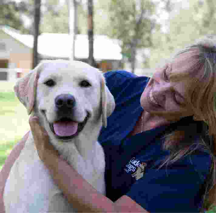 A veterinarian and fully grown yellow labrador are sitting outside. The veterinarian is looking at the dog with a smile on their face and the dog is smiling at the camera.