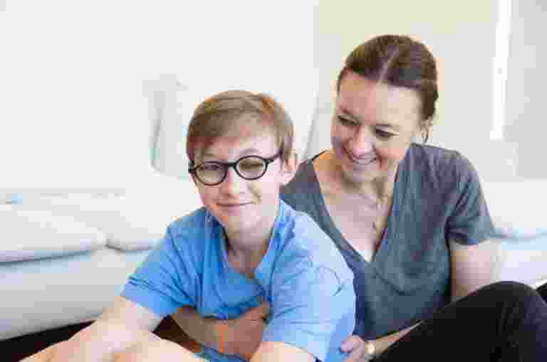 A young child with their mother seated on the ground. The mother is holding the child from behind and looking at her child. The child is looking at the camera smiling.